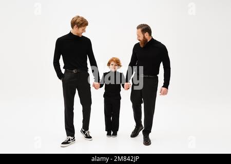 full length of happy redhead kid holding hands with bearded grandfather and dad wearing black clothes on light grey background,stock image Stock Photo