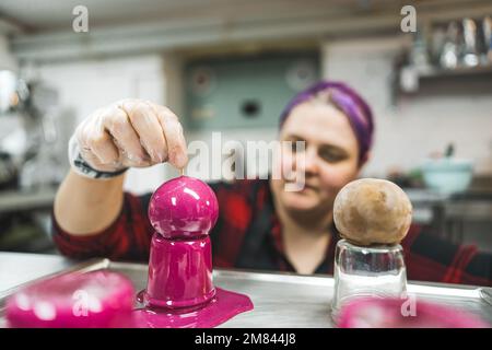 female baker with purple hair decorating chocolate balls with pink cream and putting a stick into it. High quality photo Stock Photo