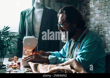Tailor man working on sewing machine in workshop with dummy wearing suit in background. Mature happy man work indoor and smile. Concept of alternative Stock Photo