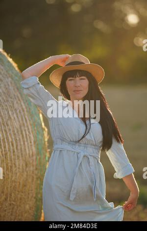 Ukrainian girl in national clothes with black thick hair and a light dress holds one hand on a straw hat on her head Stock Photo