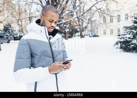 Young African American man in casual white clothes typing message, texting on phone, in park or street outdoor. Winter season with snow. Side view, copy space Stock Photo