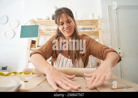 Female potter's hands roll out clay on the table with a wooden rolling pin in a craft class. Stock Photo