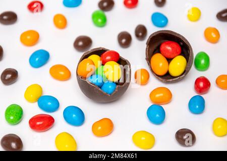 Half of a cracked chocolate egg filled with multi-colored candies on a white background with multi-colored candies, top view. Treats for children. Swe Stock Photo