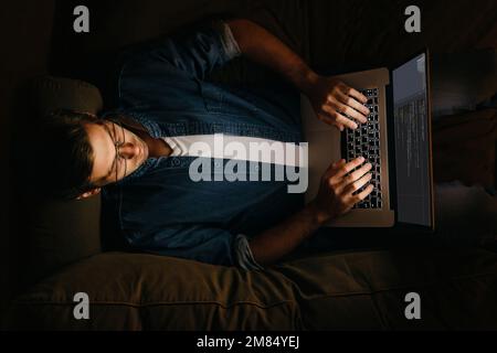 Top view of male programmer laying on sofa and working at his laptop. Overworked coder working from home at night.  Stock Photo