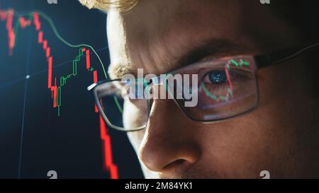 Close-up portrait of focused crypto trader analyst wearing eyeglasses looking at computer screen analyzing stock market charts. Eyeglasses reflection cryptocurrency downtrend charts. Bitcoin crash Stock Photo