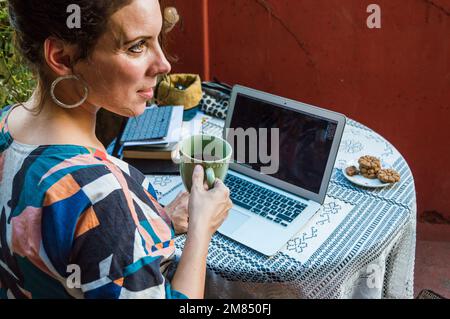 rear view busy adult woman sitting working with her laptop at the terrace drinking coffee and looking to the side, taking a break from work, lifestyle Stock Photo