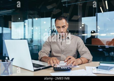 Serious thinking hispanic businessman working with documents and invoices inside office, man behind paper work, using laptop at work sitting at desk. Stock Photo