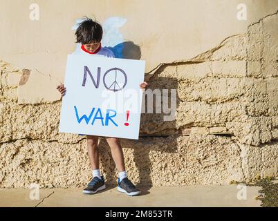 a black-haired asian boy in front of a wall, holding a sign with his hands that says "no war". the boy looks at the sign. concept stop war and peace. Stock Photo