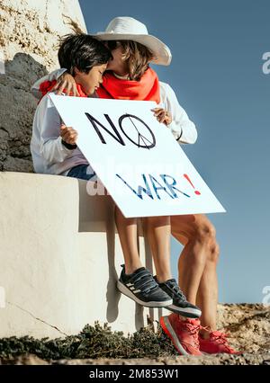 A mother comforts her Asian son in front of a bunker, while holding a sign that reads 'no to war'. concept activism for Peace in the world. Stock Photo