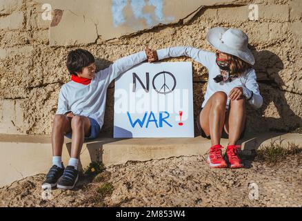 a mother and her asian son are sitting in front of a bunker and in the middle of them a sign that says 'no war'. they are holding hands. concept stop Stock Photo