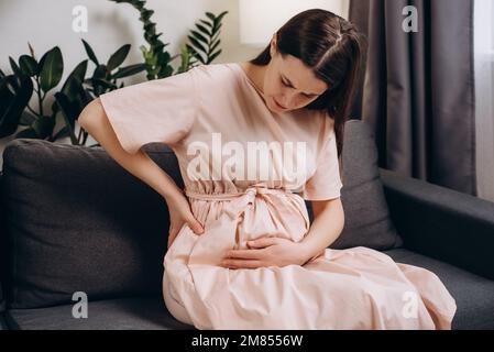 Pregnant lady having backache at home. Unhappy woman suffering from lower back pain sitting on sofa in living room. Tired pregnant female with third t Stock Photo