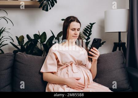 Relaxed peaceful smiling young woman with big belly sitting on cozy sofa and using modern smartphone. Pregnant lady laughing while watching some funny Stock Photo