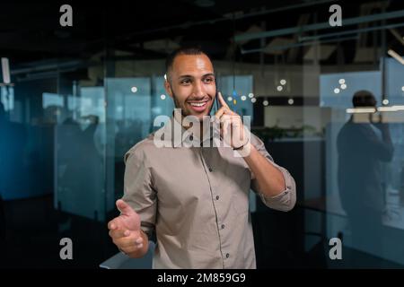 Cheerful and smiling businessman in a shirt talking on the phone inside the office, hispanic man standing near the window in the evening. Stock Photo