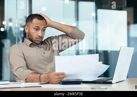 Pensive serious businessman reading financial report, hispanic businessman holding document in hands looking disappointed, working inside modern office with laptop behind paper work. Stock Photo