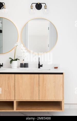 A beautiful bathroom with a floating wood cabinet, marble countertop, and gold circular mirrors. Stock Photo