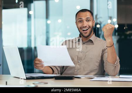 Portrait of successful hispanic businessman, man in shirt reading report and happy celebrating success satisfied with result holding hand up, worker inside office with laptop working on paper work. Stock Photo