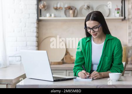 A young woman journalist, writer, blogger works remotely from home using a laptop online. Sitting in the kitchen at the table, making notes in a notebook. Stock Photo