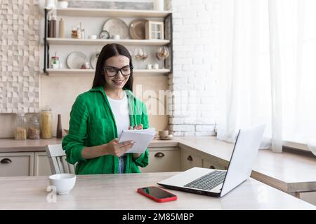 A young female journalist, writer, blogger works remotely from home using a laptop online. Sitting in the kitchen at the table, making notes in a notebook. Stock Photo