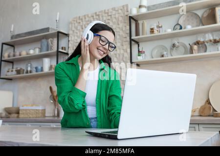 Young beautiful woman sitting in headphones at home at kitchen table and using laptop. Listens to music, enjoys, rests, smiles. Stock Photo