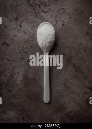 One spoonful of white refined sugar isolated on a dark kitchen counter. Top view flat lay with vertical orientation Stock Photo