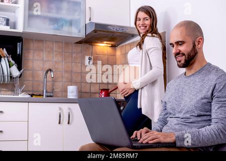 A young pregnant woman making coffee in the kitchen, while her husband is relaxing on the sofa, working on a laptop computer. Stock Photo