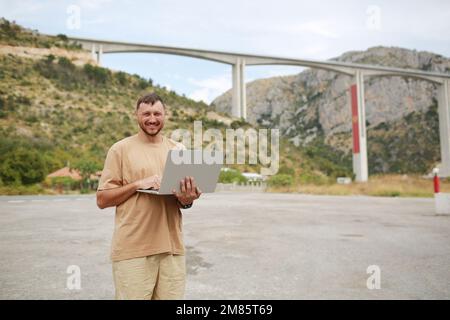 Collected man in casual clothes working on laptop outdoors on bridge construction background. Concept of remote work engineer or freelancer lifestyle. Cellular network broadband coverage. internet 5G. Stock Photo