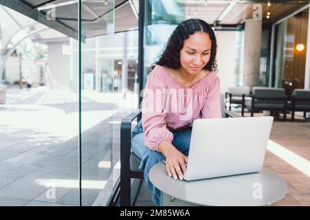 Happy young latin woman using a computer and sitting outside a cafe in her free time. Digital nomad Stock Photo