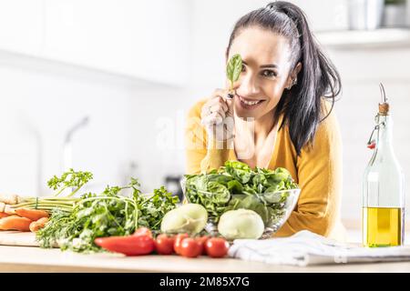 Nice young woman holding a spinach leaf, covering her eye, smiling, fresh vegetables on the table next to her. Stock Photo