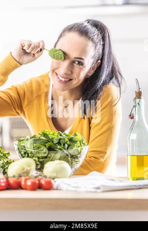 Nice young woman holding a spinach leaf, covering her eye, smiling, fresh vegetables on the table next to her. Stock Photo