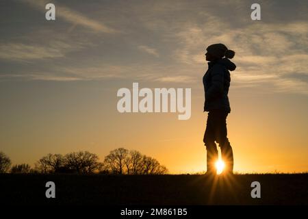 A sunburst breaks through the silhouette of an isolated woman, dressed in warm coat & bobble hat on a late afternoon, winter walk in UK countryside.