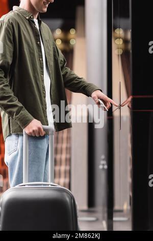 partial view of smiling traveler with baggage and smartphone standing near doors of elevator and pressing call button,stock image Stock Photo