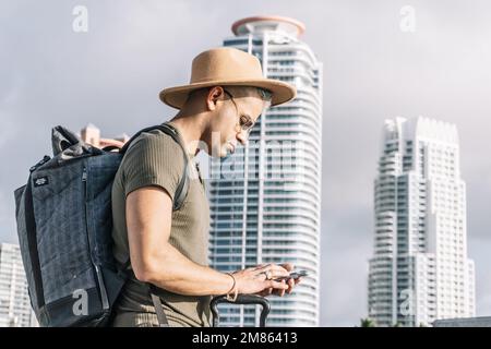 Close up, profile of traveller man with a brown hat, looking for direction on his smartphone. A skyscraper background. Stock Photo
