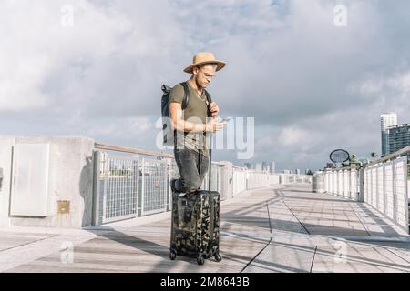 Portrait of a young man traveling with suitcase and using his smartphone. Stock Photo