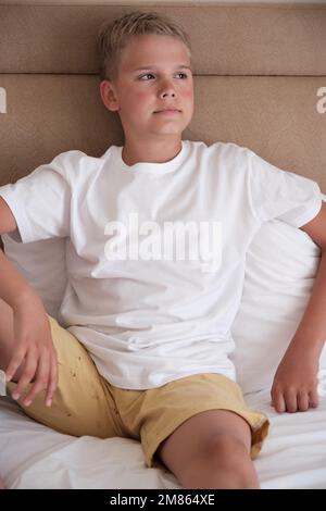 A cute young teenager sits in bed in a white t-shirt and looks out the window. Stock Photo