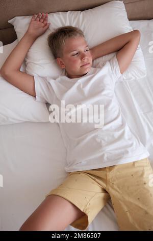 Handsome teenager lies in bed in a white t-shirt in the bedroom at home and looks out the window. Stock Photo