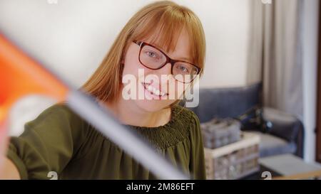 caucasian woman with ginger hair wearing glasses smiling and cleaning the camera with a squegee, house cleaning concept. High quality photo Stock Photo