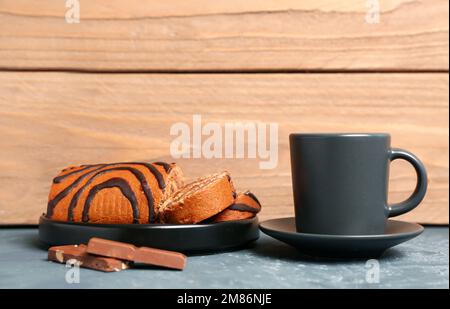 Plate with pieces of delicious sponge cake roll, cup and chocolate on grey table near wooden wall Stock Photo