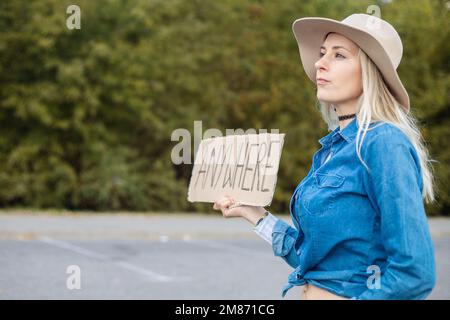 Tired woman hopefully look out passing cars with cardboard poster on roadside in forest. Lady in hat and denim outfit escape from city to go anywhere