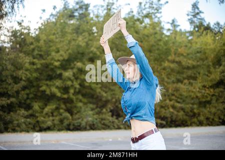 Young woman actively look out passing cars with cardboard poster on roadside in forest. Lady in hat and denim outfit escape from city to go anywhere