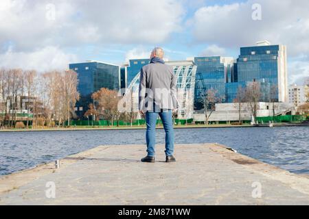 Rearview of gray haired businessman in warm jacket and scarf standing on river bridge,look office buildings. Urban area Stock Photo