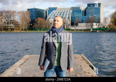 Concentrated pensive businessman with laptop in warm clothes stand on river pier nearmultistory buildings. Urban sprawl Stock Photo