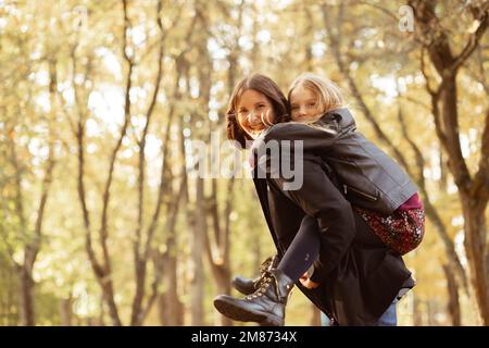 Playful, satisfied positive family of mother hold little blond girl, daughter on back, walking in golden autumn woodland Stock Photo