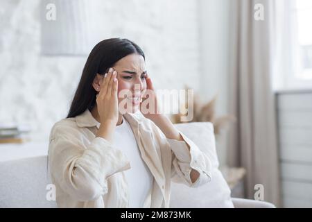 Worried young woman suffering from depression, mental disorders. He is sitting in the hospital room on the sofa near the window, holding his head with his hands, feeling a strong headache, pressure. Stock Photo