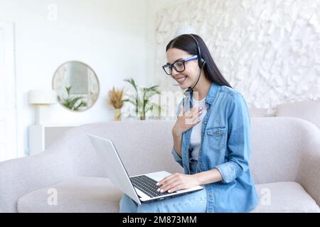 A young beautiful woman is having a remote job interview online. Sitting at home on the sofa with a laptop in a headset. Talking via video call. Stock Photo