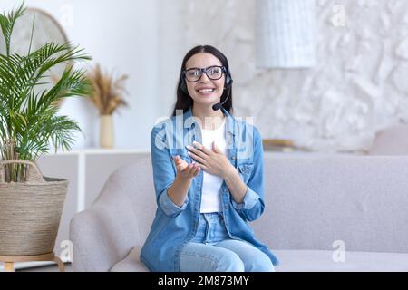 A young woman is having a remote job interview online. Sitting at home on the sofa in a headset. Talks through a video call, introduces himself, looks into the camera, smiles. Stock Photo