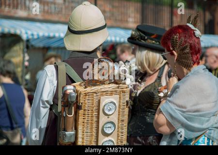 A group of people at a steampunk festival. A man in a pith helmet has a wicker basket with dials on his back and a cute toy dog sitting on it. Stock Photo