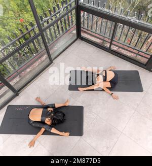 Two attractive women doing yoga kurmasana posture in studio, on the floor, wearing sportswear. Stock Photo