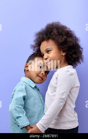 two pretty kid friends hugging holding hands together isolated over purple pastel color background, adorable kid boy and girl in casual wear posing ha Stock Photo