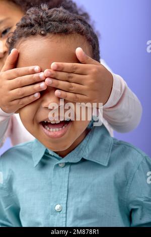 Happy little girl have fun with brother boy 5-6 years old, close eyes with hands play guess who isolated on purple background in studio. focus on laug Stock Photo