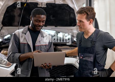Handsome diverse mechanics in uniform examining car, using laptop while working in modern auto service. black and caucasian Auto mechanics checking ca Stock Photo
